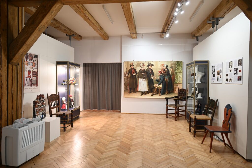 Przeszklony regał z wystawionymi elementami kultury jamneńskiej. Po prawej i po lewej - krzesła z polichromią jamneńską. Po środku współczesny wydruk litografii ze sceną (kliknięcie spowoduje powiększenie zdjęcia).