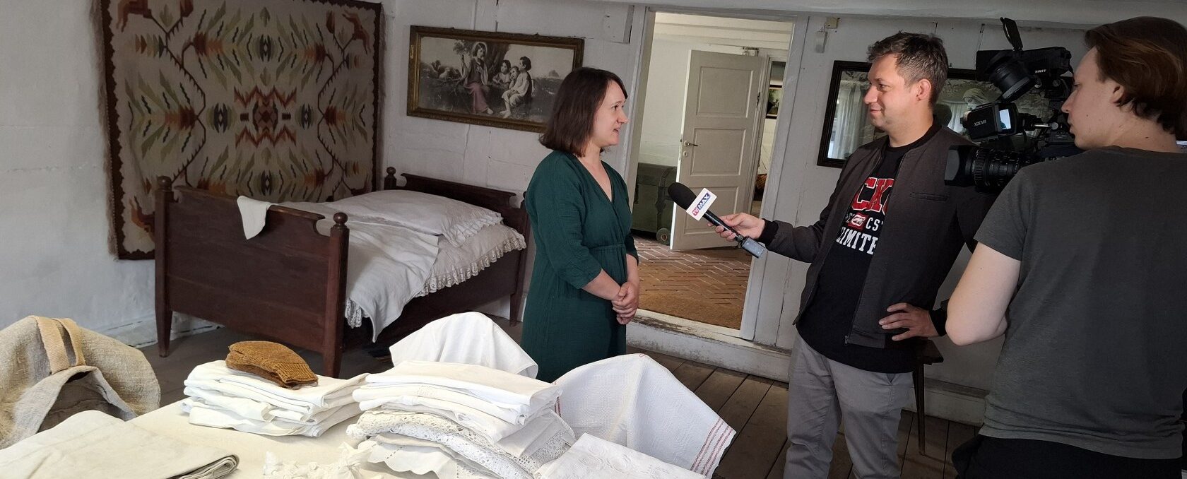 Kobieta udziela wywiadu dla telewizji. Trzy osoby stoją w starej chacie. W tle meble z epoki.