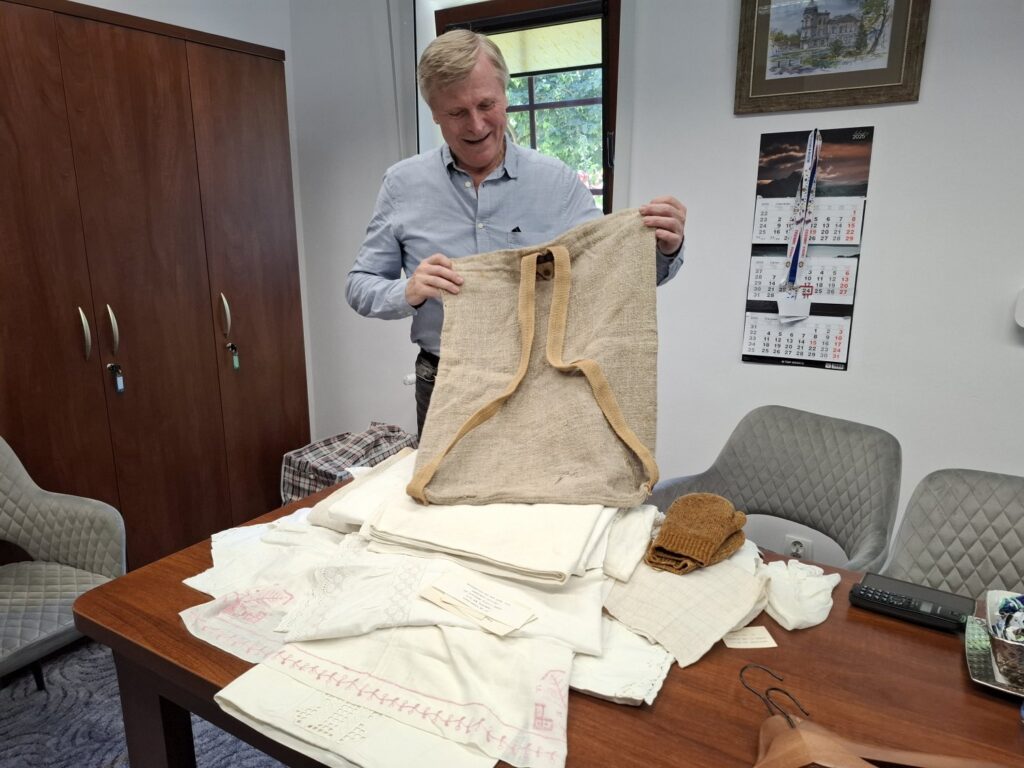 Mężczyzna trzyma w rękach artefakt przekazany do muzeum. Na stole leżą materiały tekstylne. W tle widoczna szafa (kliknięcie spowoduje powiększenie zdjęcia).