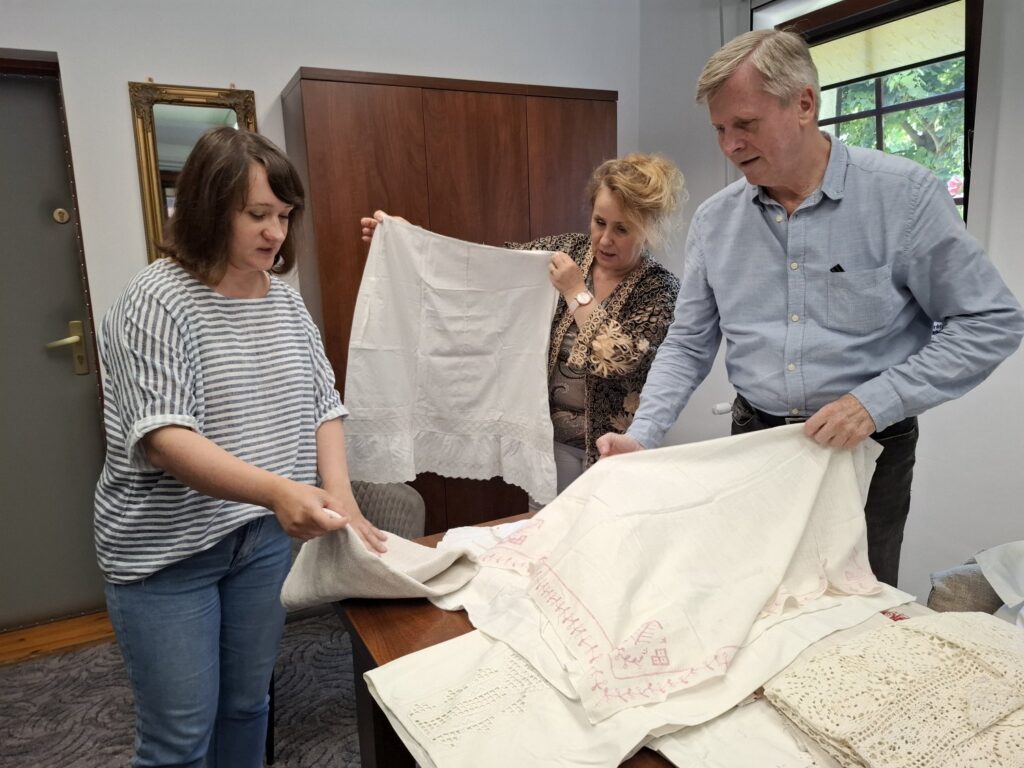 Trzy osoby pokazują do obiektywu artefakty przekazane do muzeum - odzież i tekstylia (kliknięcie spowoduje powiększenie zdjęcia).