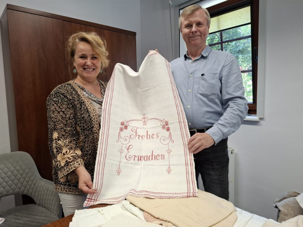 Dwie osoby trzymają w rękach ręcznie haftowaną makatkę z napisem "Frohes Erwachen" - miłego przebudzenia (kliknięcie spowoduje powiększenie zdjęcia).