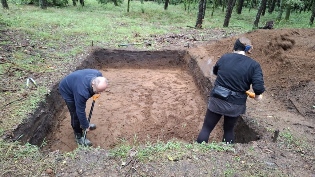 Wykop archeologiczny w lesie, w którym pracują dwie osoby (kliknięcie spowoduje powiększenie zdjęcia).