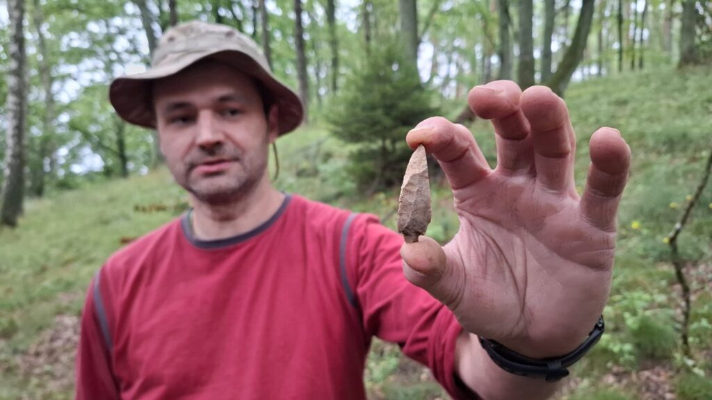 Archeolog prezentuje, trzymany w dłoni, grot włóczni z krzemienia. W tle porośnięte drzewami wzgórze (kliknięcie spowoduje powiększenie zdjęcia).