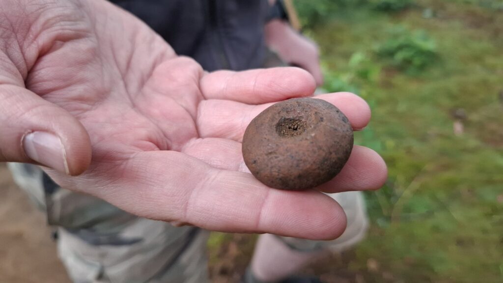 Archeolog prezentuje, trzymany na dłoni, ciężarek tkacki (kliknięcie spowoduje powiększenie zdjęcia).