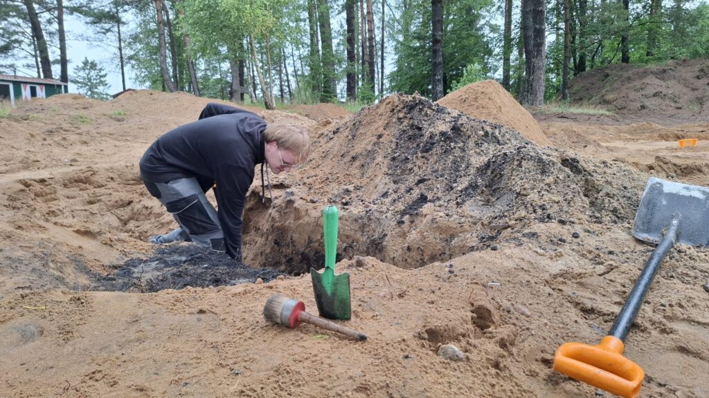Wolontariusz pogłębia wykop archeologiczny. W tle las. Na pierwszym planie narzędzia: pędzel, mała łopatka i łopata (kliknięcie spowoduje powiększenie zdjęcia).