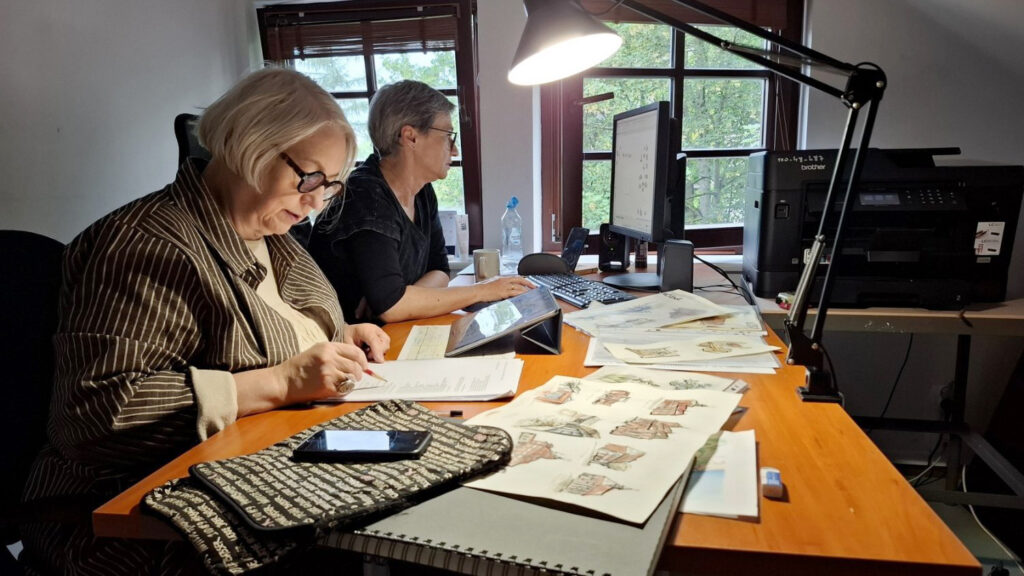 Dwie kobiety siedzą przed biurkiem, na którym rozłożone są projekty rysunków różnych obiektów architektonicznych (kliknięcie spowoduje powiększenie zdjęcia).