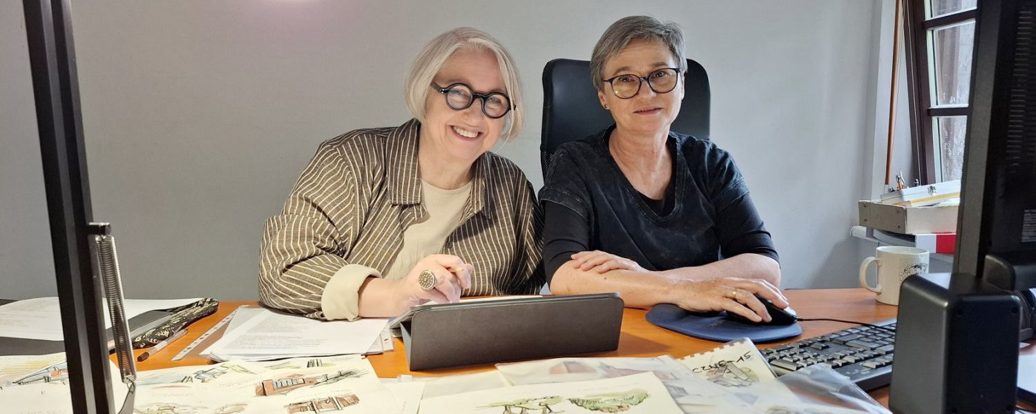 Dwie kobiety siedzą przed biurkiem, na którym rozłożone są projekty rysunków różnych obiektów architektonicznych.