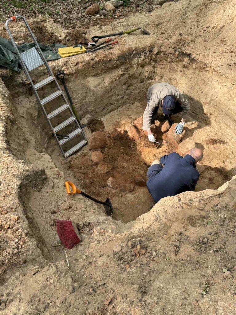 Dwie osoby siedzą w wykopalisku archeologicznych i odsłaniają fragment stanowiska archeologicznego (kliknięcie spowoduje powiększenie zdjęcia).