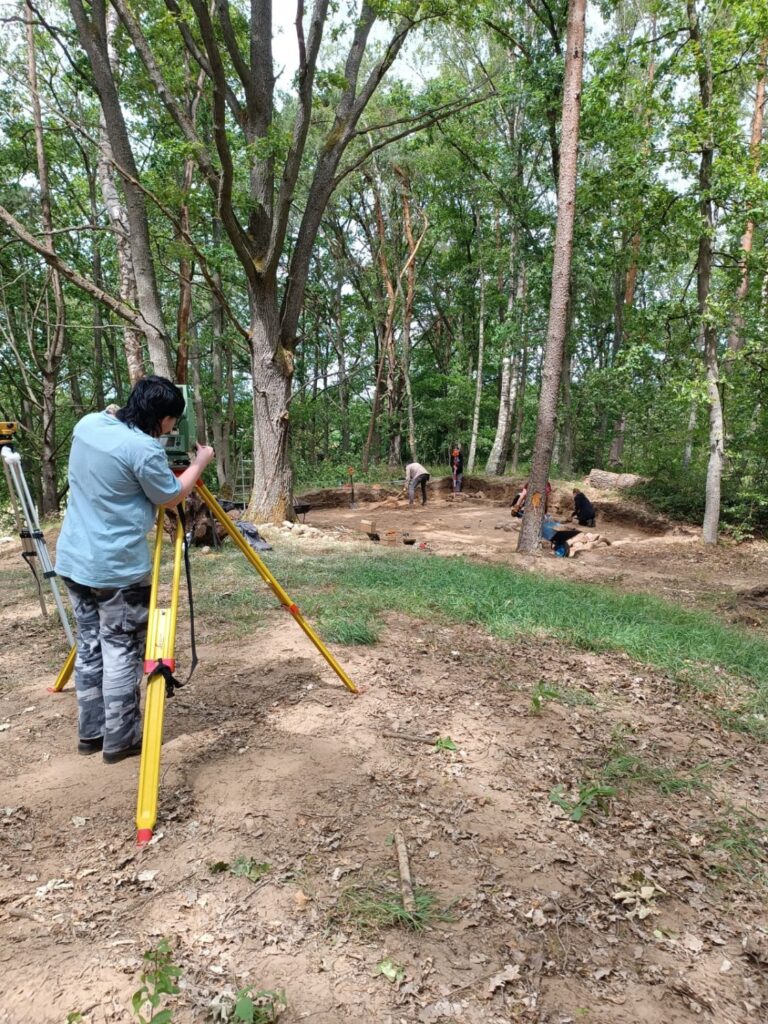 Osoba stoi przy urządzeniu mierniczym. W tle wykop archeologiczny, w którym pracują ludzie (kliknięcie spowoduje powiększenie zdjęcia).