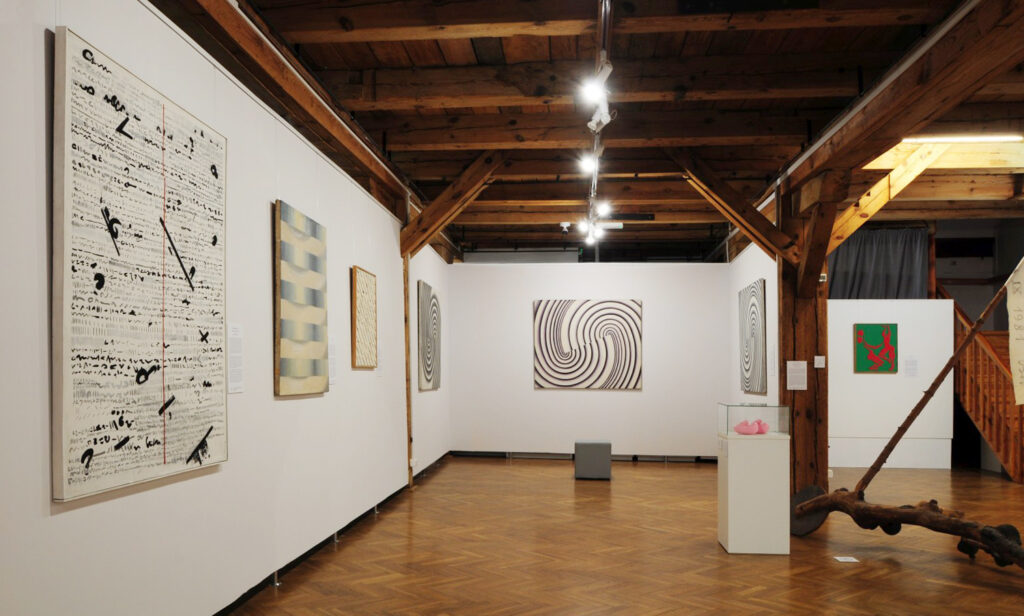 Na ściankach wystawowych wiszą obrazy, po środku pufa, a po prawej praca pod tytułem Taczki wolności - kliknij aby powiększyć zdjęcie.