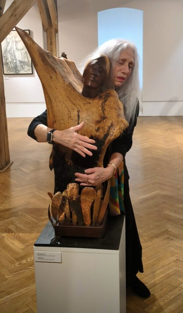 Kobieta stoi na sali wystawowej i obejmuje rzeźbę stojącą na podeście (kliknięcie spowoduje powiększenie zdjęcia).