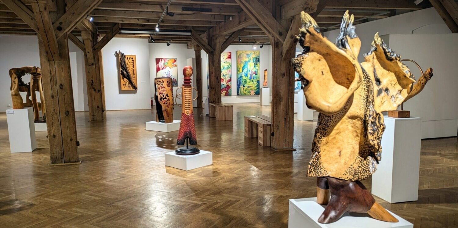 Sala wystawowa, na której znajdują się drewniane rzeźby ułożone na podestach. W tle obrazy powieszone na ścianach.