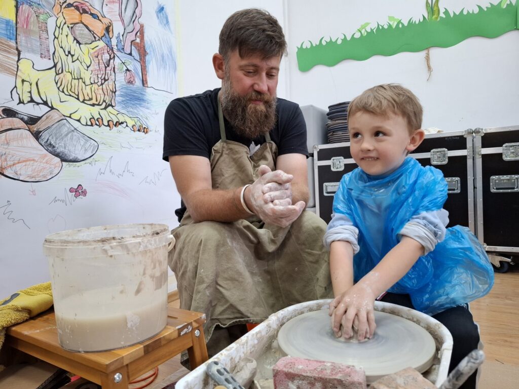 Chłopiec, w obecności dorosłego prowadzącego warsztaty, siedzi przy kole garncarskim i obrabia formę z gliny (kliknięcie spowoduje powiększenie zdjęcia).