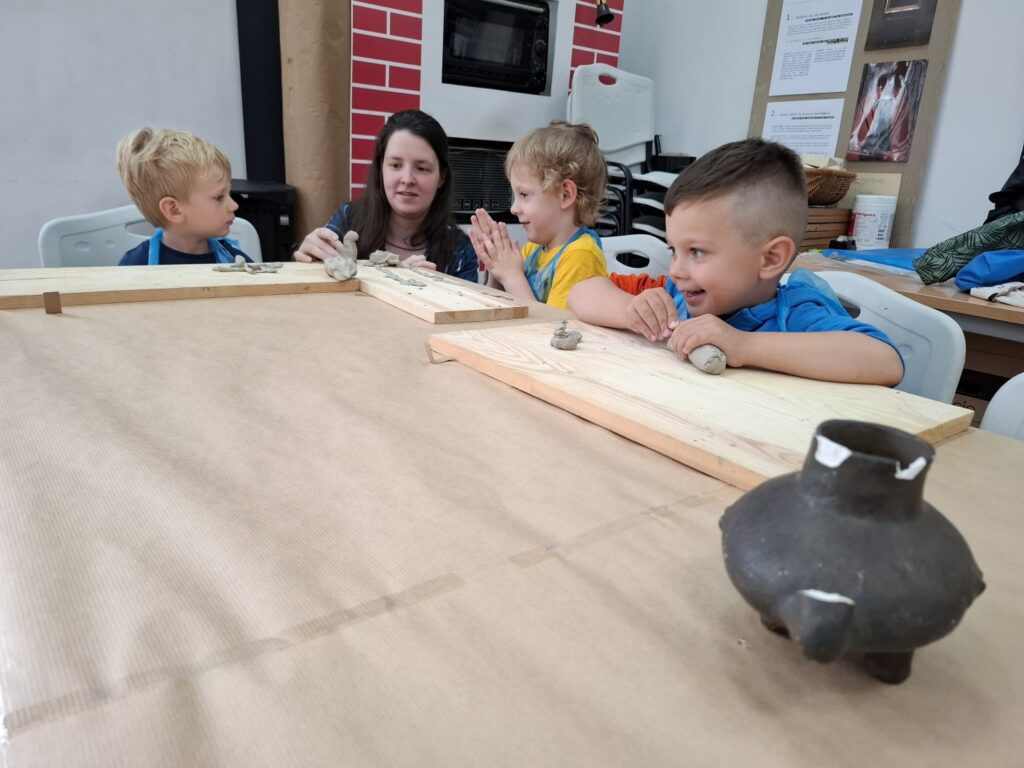 Trójka dzieci, wraz z prowadzącą zajęcia, siedzi przy stole i modeluje przedmioty z gliny. Na pierwszym planie, gliniana rekonstrukcja dawnego naczynia (kliknięcie spowoduje powiększenie zdjęcia).