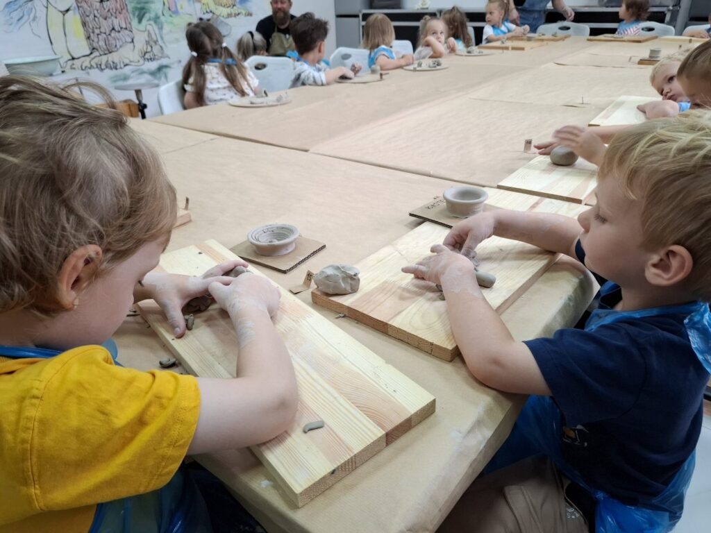 Dzieci siedzą przy stole i modelują przedmioty z gliny. (kliknięcie spowoduje powiększenie zdjęcia).