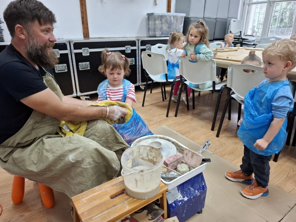 Pięcioro dzieci obserwuje osobę prowadzącą zajęcia i pokazującą, jak należy pracować z kołem garncarskim (kliknięcie spowoduje powiększenie zdjęcia).