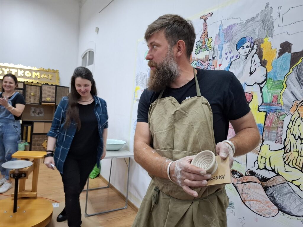 Prowadzący zajęcia, pokazuje grupie dzieci, wykonane z gliny, małe naczynie. W tle salka edukacyjna i dwie panie prowadzące zajęcia (kliknięcie spowoduje powiększenie zdjęcia).