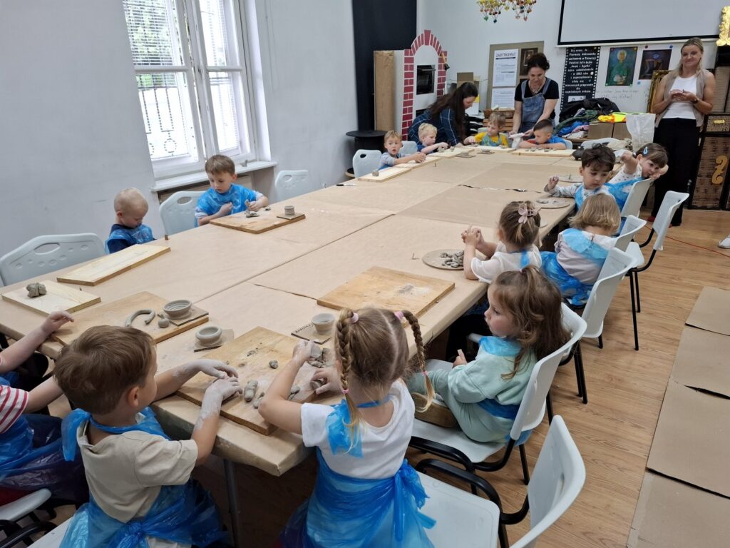 Dzieci siedzą przy stołach rozstawionych na sali edukacyjnej i lepią naczynia z gliny (kliknięcie spowoduje powiększenie zdjęcia).