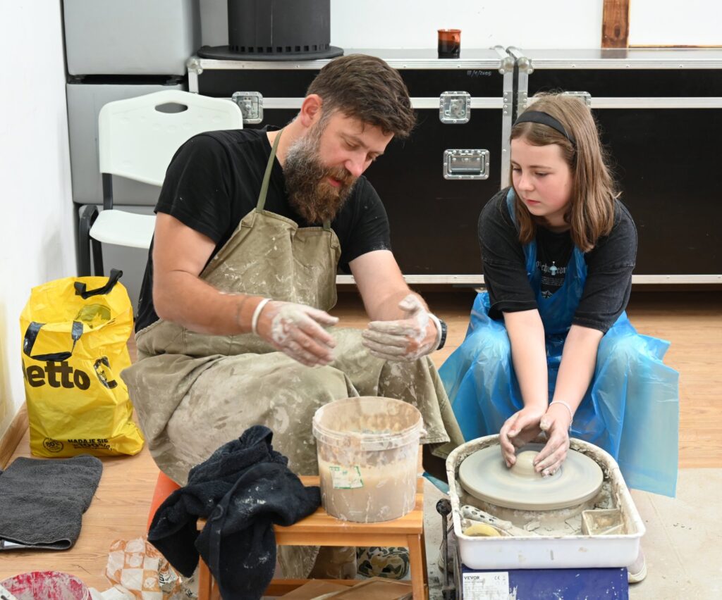Prowadzący zajęcia z garncarstwa, pokazuje dziewczynce, jak należy pracować przy kole garncarskim. Obydwie osoby siedzą przed kołem na taboretach (kliknięcie spowoduje powiększenie zdjęcia).