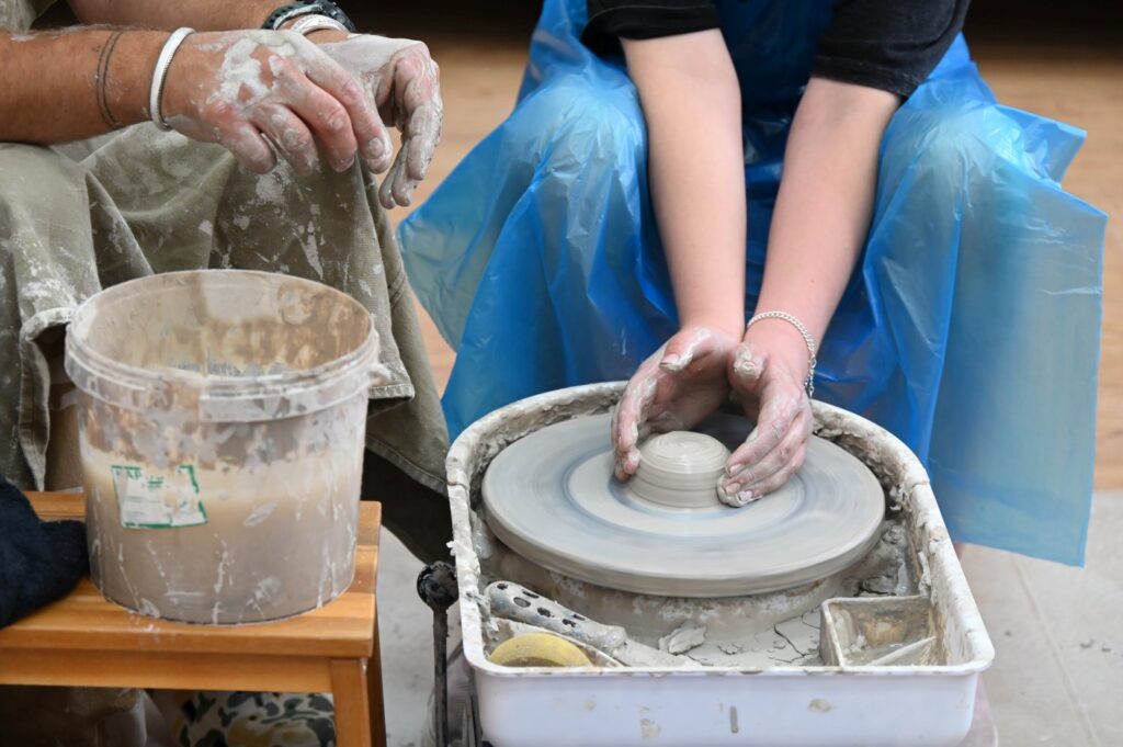 Zbliżenie na koło garncarskie i dłonie osoby, który przy nim pracuje i formuje jakiś przedmiot z gliny (kliknięcie spowoduje powiększenie zdjęcia).