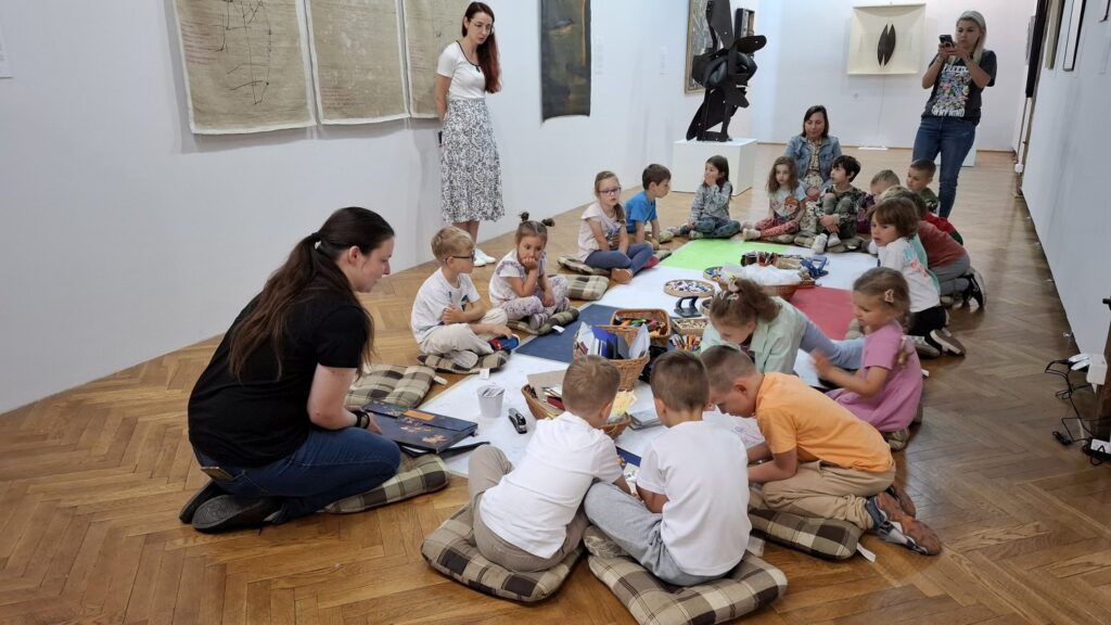 Grupa dzieci siedzi na poduszkach, na podłodzie i szykuje się do malowania artystycznej książeczki. W tle fragmenty wystawy sztuki współczesnej (kliknięcie spowoduje powiększenie zdjęcia).