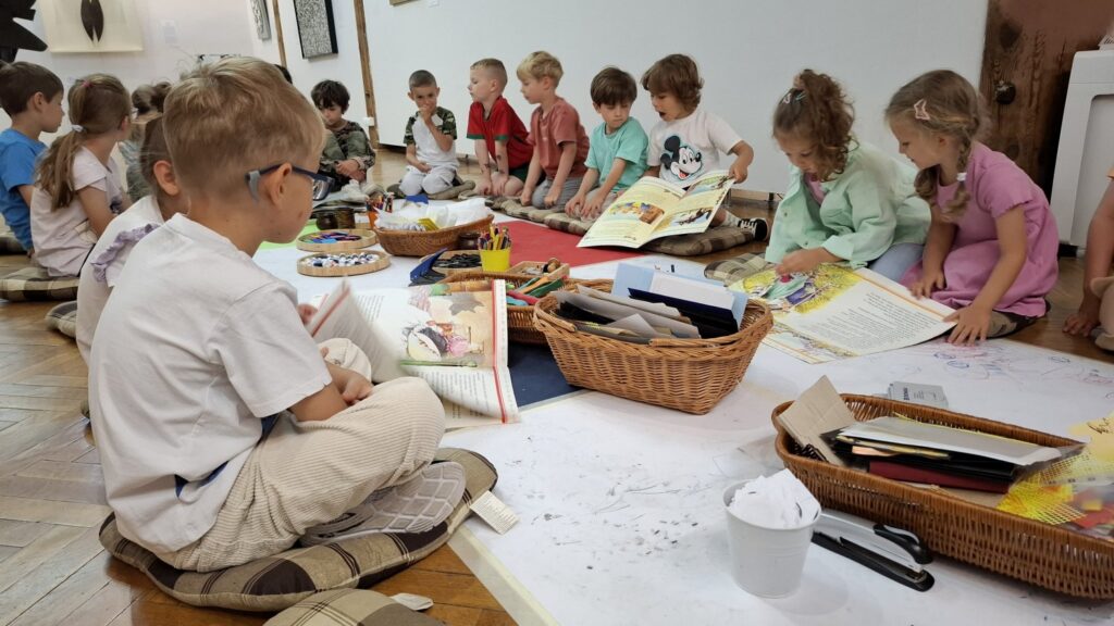 Siedzące na podłodze dzieci, szykują się do malowania artystycznej książeczki. Przed nimi kartki, kredki i farbki (kliknięcie spowoduje powiększenie zdjęcia).