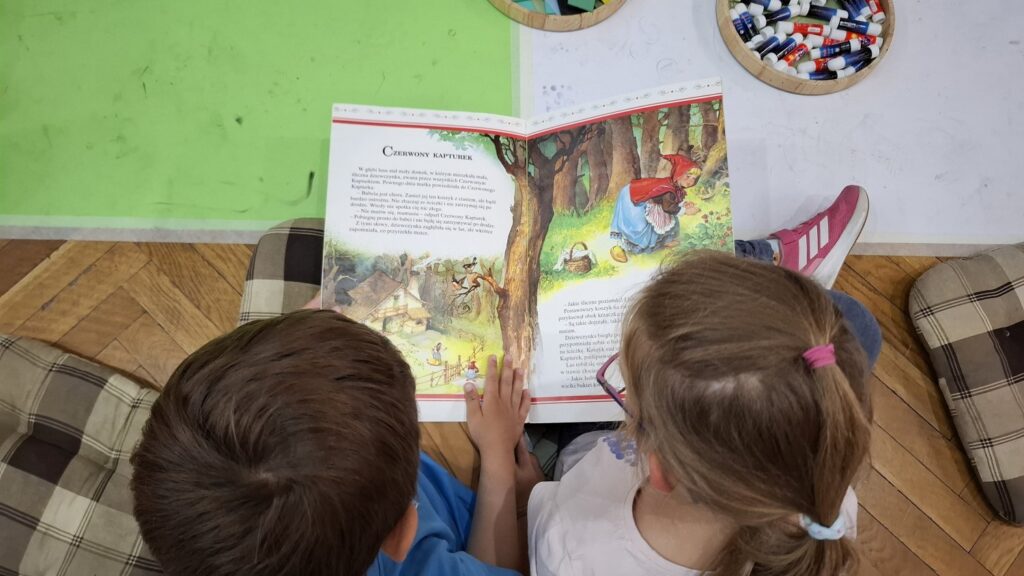 Dwoje dzieci siedzi na podłodze, trzyma w rękach książkę i czyta bajkę o czerwonym kapturku (kliknięcie spowoduje powiększenie zdjęcia).