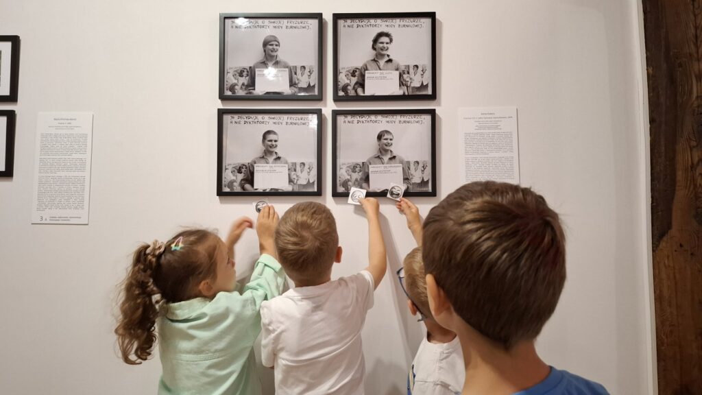 Dzieci stoją przed zawieszonymi na ścianie czterema zdjęciami i pokazują namalowane podczas warsztatów emotikonki (kliknięcie spowoduje powiększenie zdjęcia).