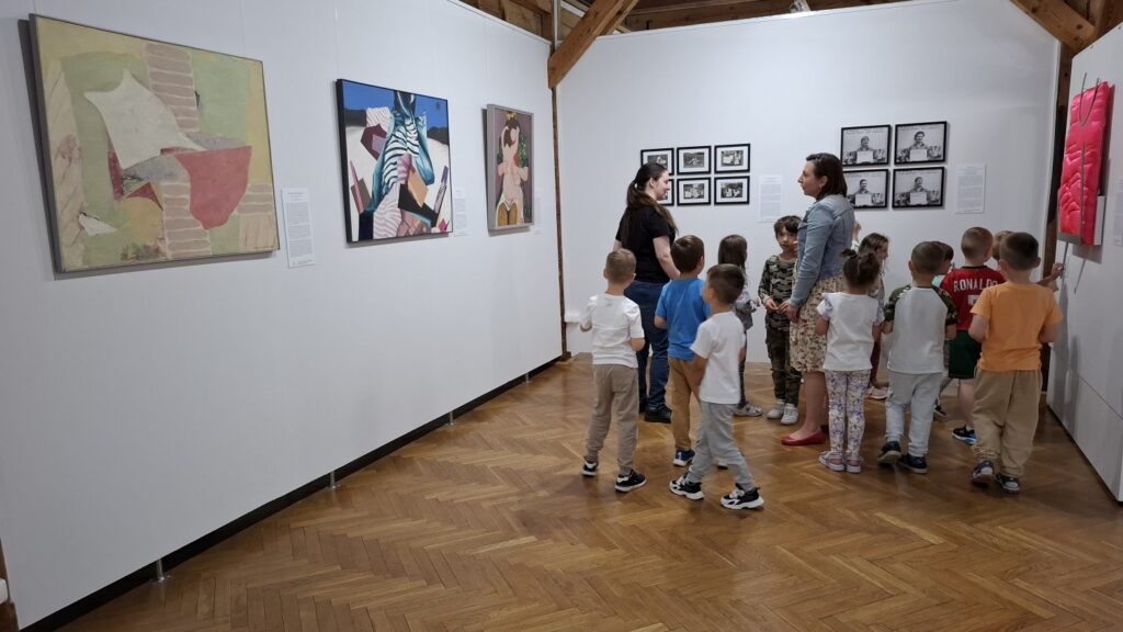 Grupa dzieci, wraz z opiekunami, zwiedza wystawę sztuki współczesnej. Na ścianach wiszą obrazy i zdjęcia (kliknięcie spowoduje powiększenie zdjęcia).