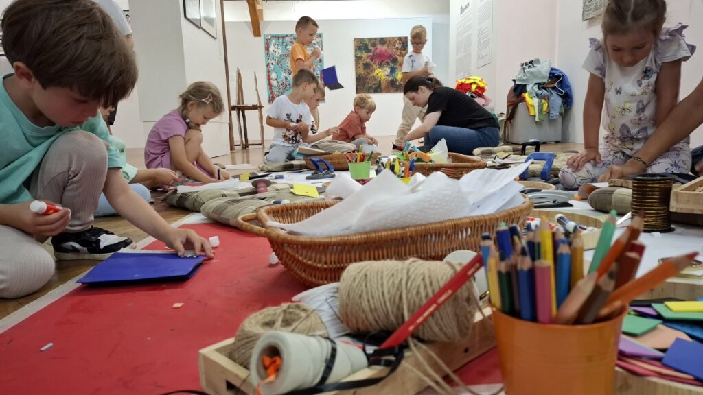 Siedzące na podłodze dzieci malują artystyczną książeczkę. w tle widoczne obrazy z wystawy sztuki dawnej (kliknięcie spowoduje powiększenie zdjęcia).