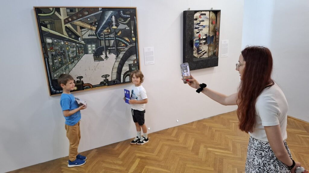 Kobieta fotografuje dwójkę chłopców stojących przed obrazem wystawy sztuki współczesnej. W ręce trzymają wykonane przez siebie książeczki artystyczne (kliknięcie spowoduje powiększenie zdjęcia).
