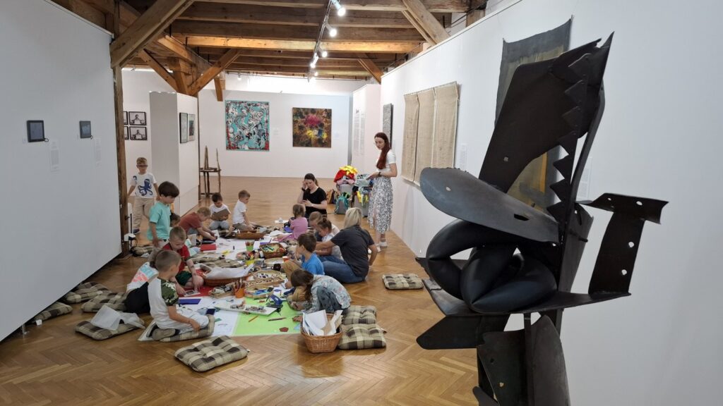 Siedzące na podłodze dzieci malują artystyczną książeczkę. w tle widoczne obrazy z wystawy sztuki dawnej, a na pierwszym planie wykonana z metalu rzeźba (kliknięcie spowoduje powiększenie zdjęcia).