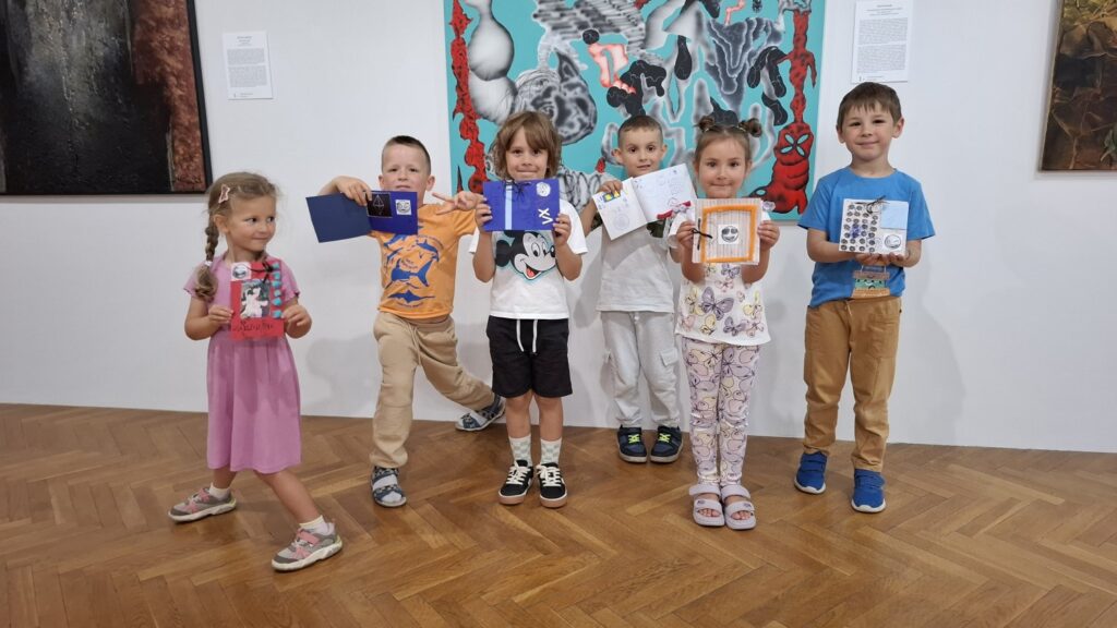 Grupka dzieci stoi przed obrazem z wystawy działu sztuki współczesnej i prezentuje wykonane przez siebie książeczki artystyczne (kliknięcie spowoduje powiększenie zdjęcia).