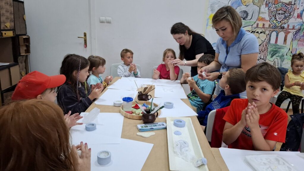 Duża grupa dzieci siedzi przy stole zastawionym kartkami i pędzlami. Uczestnicy warsztatów z reliefu (kliknięcie spowoduje powiększenie zdjęcia).