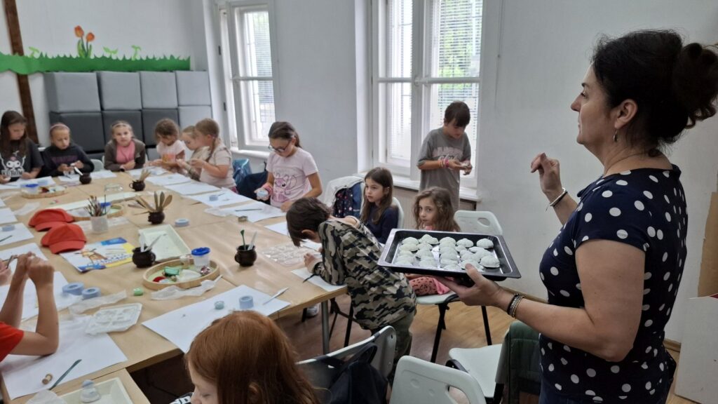 Na pierwszym planie osoba trzyma blachę z formami przygotowanymi do utwardzenia w piekarniku. W tle dzieci uczestniczące w warsztatach (kliknięcie spowoduje powiększenie zdjęcia).