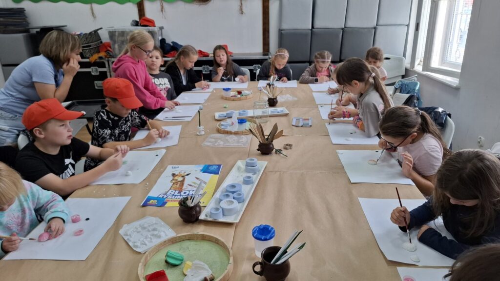 Dzieci siedzą przy stole zastawionym kartkami i pędzlami. Uczestnicy warsztatów z reliefu (kliknięcie spowoduje powiększenie zdjęcia).