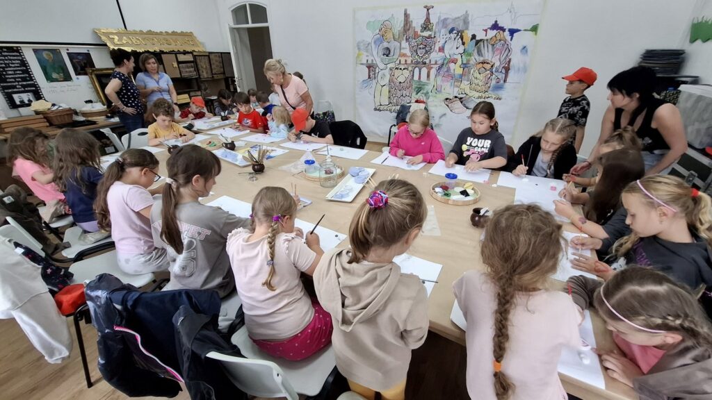 Dzieci siedzą przy stole zastawionym kartkami i pędzlami. Uczestnicy warsztatów z reliefu (kliknięcie spowoduje powiększenie zdjęcia).