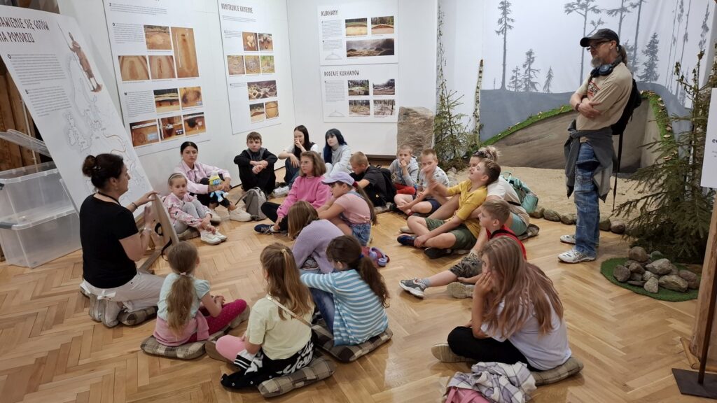 Dzieci siedzą na poduszkach, na podłodze i słuchają wykładu o gotach. W tle tablice wystawowe związane z tematyką gotów (kliknięcie spowoduje powiększenie zdjęcia).