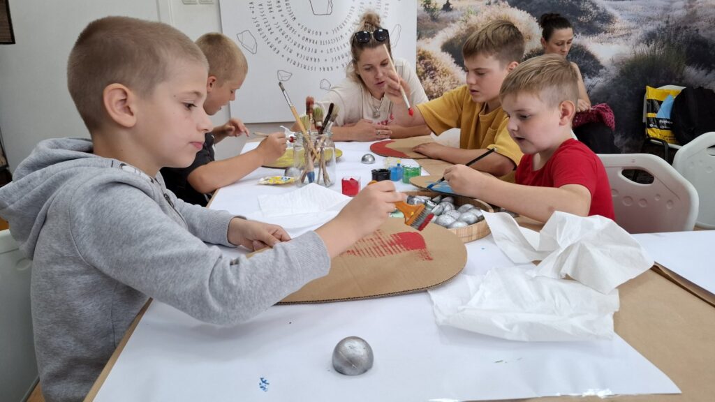 Stół z narzędziami do malowania. Przy stole siedzą dzieci biorące udział w warsztatach (kliknięcie spowoduje powiększenie zdjęcia).