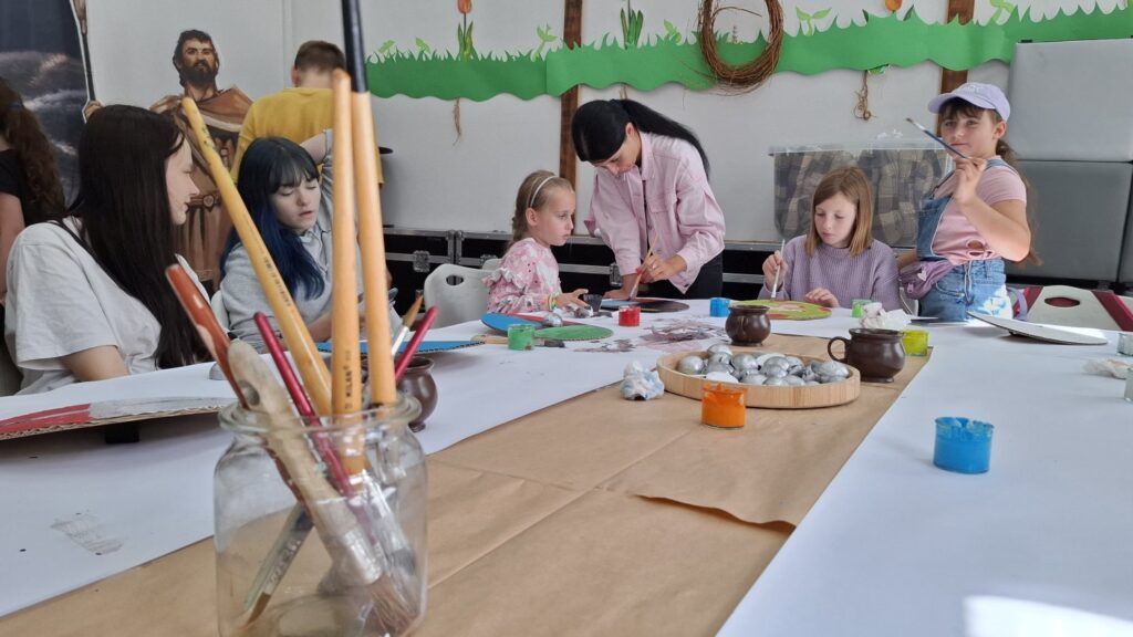 Stół z narzędziami do malowania - na pierwszym planie słoik z pędzlami. Przy stole siedzą dzieci biorące udział w warsztatach (kliknięcie spowoduje powiększenie zdjęcia).