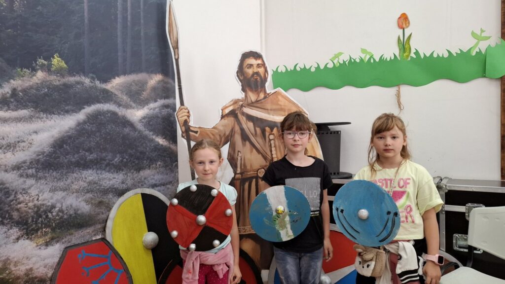 Trójka dzieci, stojących na tle figury gota, prezentuje tarcze wykonane w trakcie warsztatów (kliknięcie spowoduje powiększenie zdjęcia).