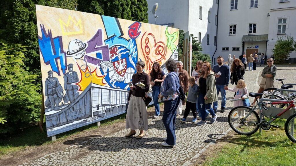 Ludzie chodzą na terenie muzeum - w tle widoczny budynek muzeum. Po lewej stronie graffiti wykonane z okazji 80 lat polskiego Koszalina. Po prawej stojaki rowerowe i rowery (kliknięcie spowoduje powiększenie zdjęcia).