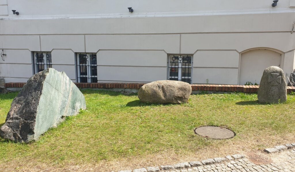 Trzy kamienie stojące obok siebie, na trawniku. W tle mury Muzeum w Koszalinie (kliknięcie spowoduje powiększenie zdjęcia).
