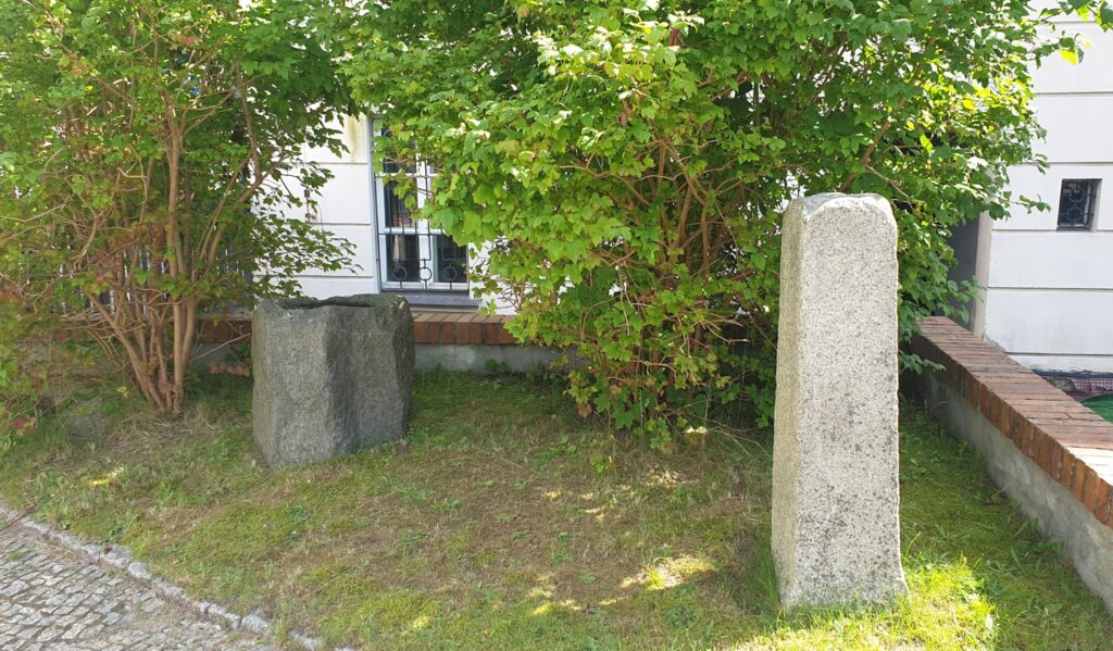 Średniowieczna chrzcielnica ustawiona pomiędzy krzewami. W tle mury Muzeum w Koszalinie (kliknięcie spowoduje powiększenie zdjęcia).