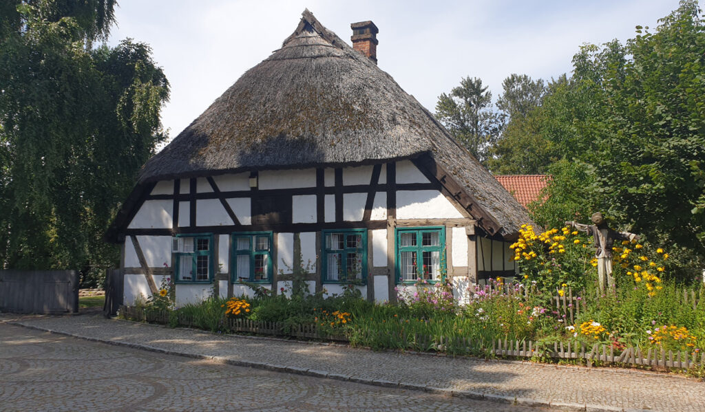 Zabytkowa chałupa rybacka z Dąbek zbudowana w technice szachulcowej. W tle widoczne drzewa, a na pierwszym planie kwietny ogród (kliknięcie spowoduje powiększenie zdjęcia).