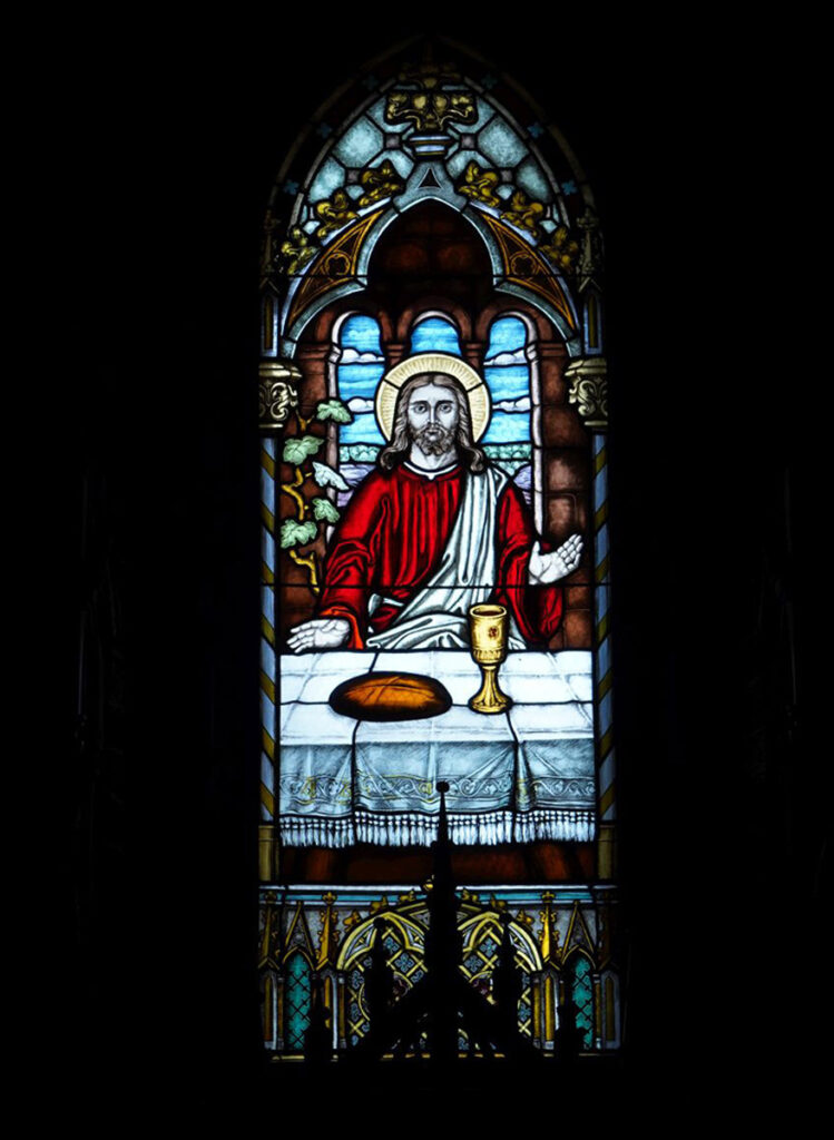 Kolorowy witraż ze sceną "Ustanowienie Eucharystii" Za stołem, na którym leży chleb i stoi złoty kielich, siedzi Jezus z wyciągniętymi w geście błogosławieństwa dłońmi (kliknięcie spowoduje powiększenie zdjęcia).