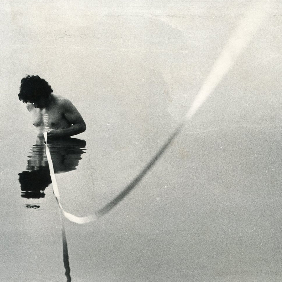 Mężczyzna stoi w jeziorze, zanurzony po pas. Ciągnie ze sobą kawałek liny. Działanie artystyczne z cyklu linia i ciało.