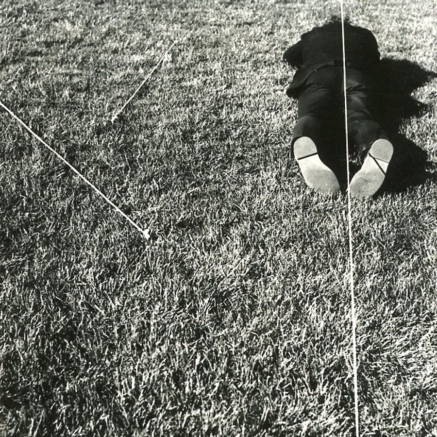 Mężczyzna leży na trawie, plecami zwróconymi w górę. Przez środek ciała przechodzi lina. Działanie artystyczne z cyklu linia i ciało.