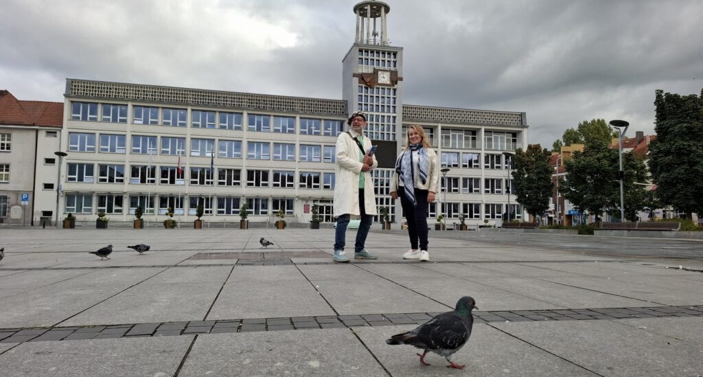 Dwie osoby stoją na tle budynku Ratusza w Koszalinie. Na pierwszym planie, po płycie rynku, chodzi gołąb.