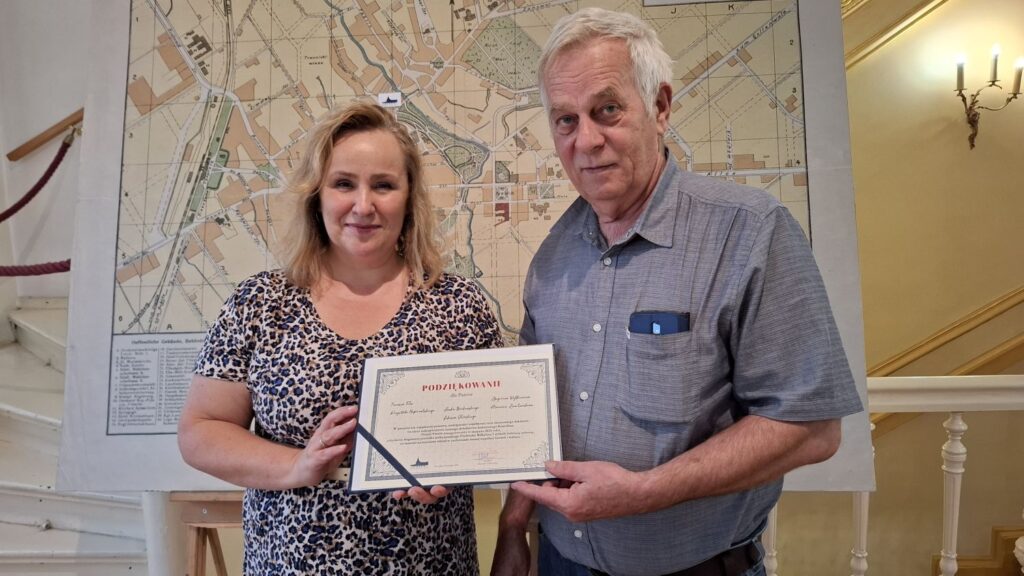 
Dwie osoby stoją przed wielkoformatową mapą Koszalina i trzymają w rękach dyplom z podziękowaniami (kliknięcie spowoduje powiększenie zdjęcia).